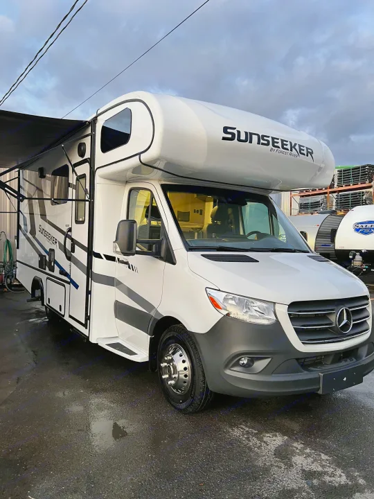 This white Sunseeker Class C RV, perfect for your next rv rental adventure, is parked with its awning out, built on a Mercedes-Benz chassis.. Forest River Sunseeker 2024