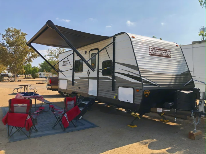 A Coleman RV rental travel trailer is set up at a sunny campsite with an extended awning, picnic table, and chairs.. Dutchmen Coleman Lantern LT 2021