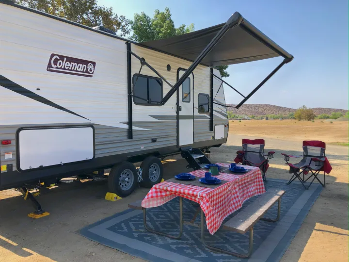 RV rental: A Coleman travel trailer parked at a campsite with an extended awning, a picnic table set with dishes, and camping chairs.. Dutchmen Coleman Lantern LT 2021