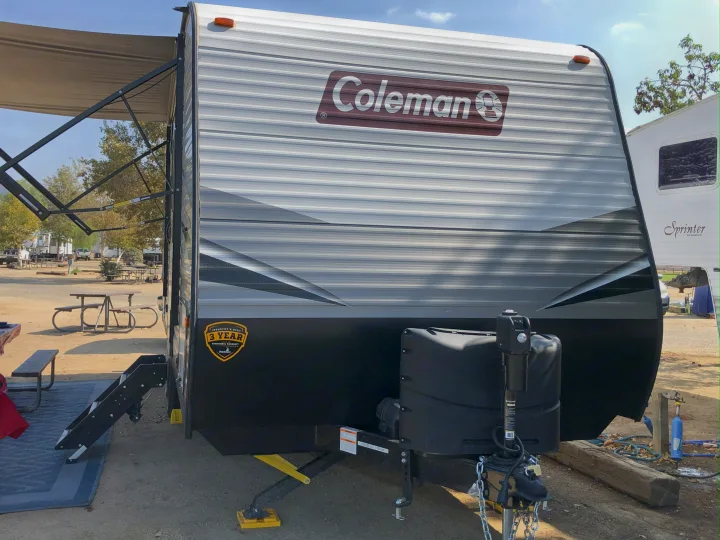 A Coleman travel trailer, ideal for an RV rental, is set up at a sunny campground with its awning extended and steps down.. Dutchmen Coleman Lantern LT 2021