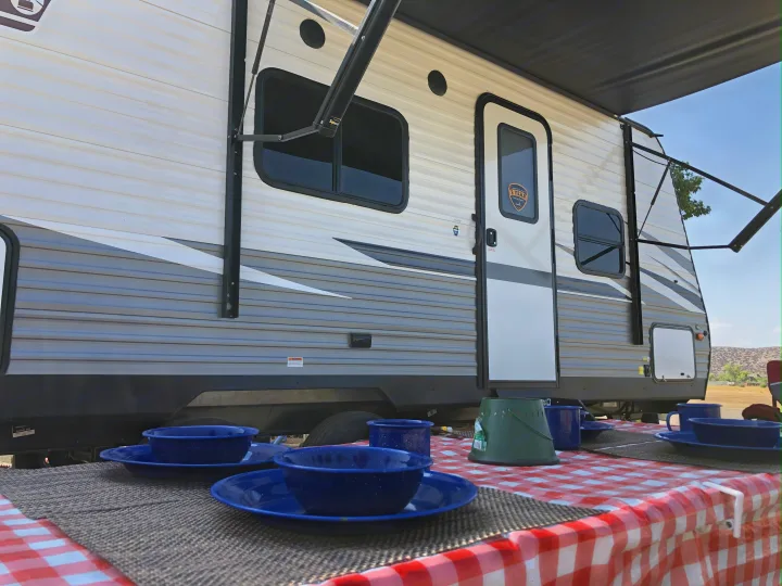 RV rental with extended awning and a picnic table set for an outdoor meal with blue enamelware, offering a comfortable campsite.. Dutchmen Coleman Lantern LT 2021