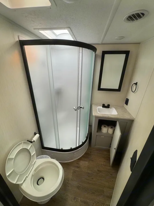 Clean RV rental bathroom featuring a curved shower stall, toilet, sink vanity with mirror, and light-colored interior.. Forest River Vibe 2021