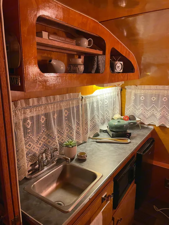 A well-appointed rv rental kitchen featuring rich wood cabinetry, a sink, microwave, induction cooktop, and lace curtains.. Play-Mor Other 1978