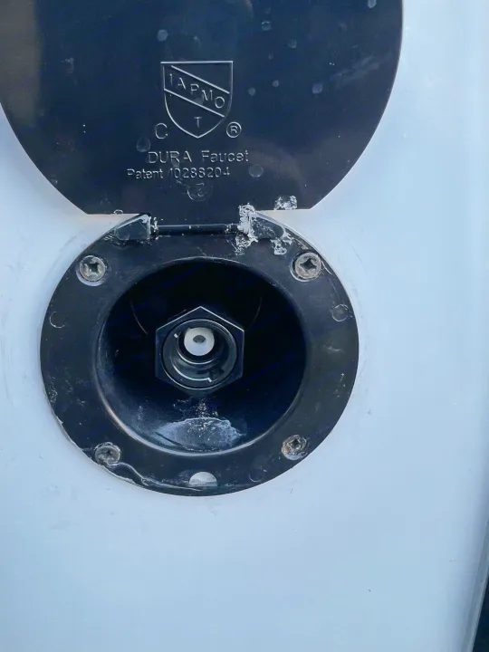 RV rental's exterior water hookup, a black Dura Faucet port with an open cover and visible connection for utilities.. Forest River Wildwood X-LITE 240BHXL 2024