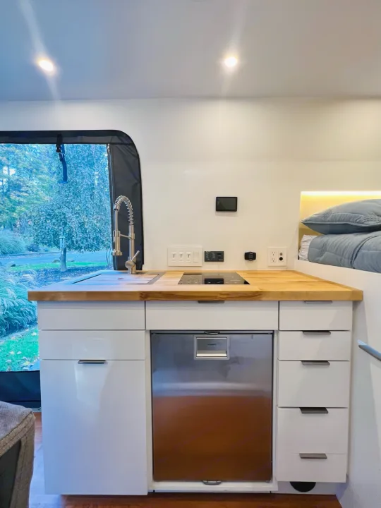 Explore this modern rv rental's interior! Features a compact kitchen with wood counter, integrated fridge, and cozy bed.. RAM Promaster 3500 extended wheel base 2023