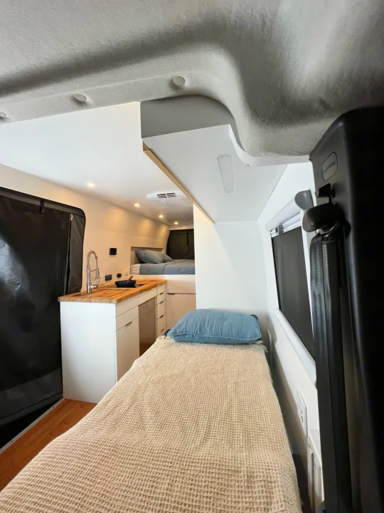 Bright RV rental interior: compact kitchen with wood counter/sink, plus two cozy bed areas visible.. RAM Promaster 3500 extended wheel base 2023