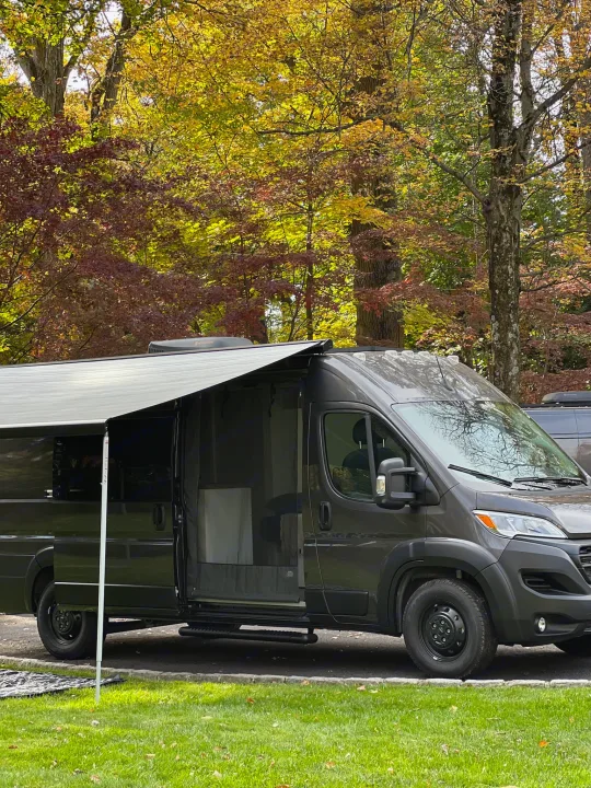 Dark grey RV rental camper van with open door and extended awning, parked among vibrant autumn trees.. RAM Promaster 3500 extended wheel base 2023