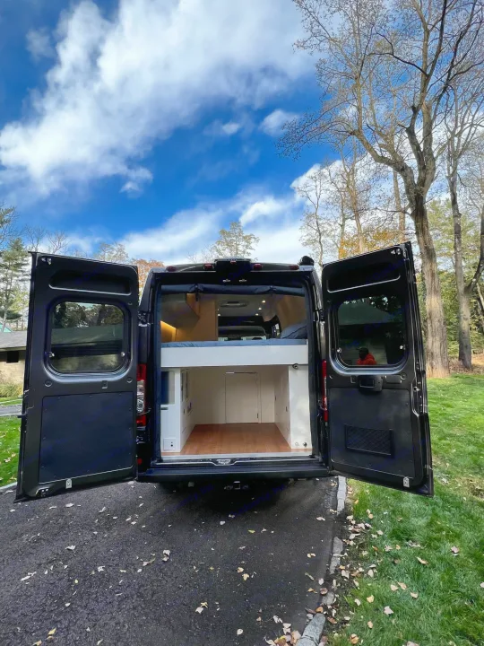This black camper van RV rental has its rear doors open, revealing a cozy interior with a bed and storage. Parked outdoors.. RAM Promaster 3500 extended wheel base 2023