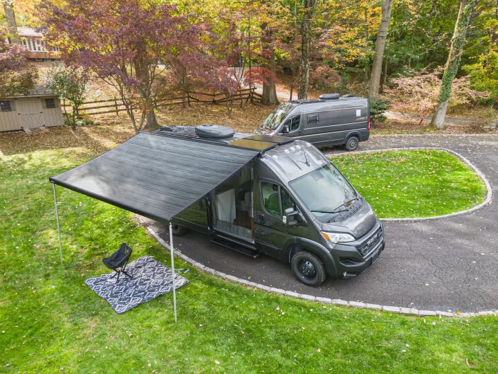 Experience adventure with an RV rental! Two dark gray campervans on a driveway, one with an extended awning, chair & blanket.. RAM Promaster 3500 extended wheel base 2023