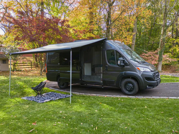 Dark gray camper van with awning extended, chair & blanket on grass in autumn. Ideal for your RV rental getaway.. RAM Promaster 3500 extended wheel base 2023