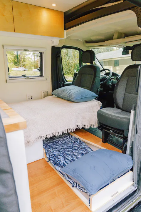 Inside a modern rv rental: a cozy bed, pull-out floor cushion, light wood cabinets, and clear view to the front cabin.. RAM Promaster 3500 extended wheel base 2023