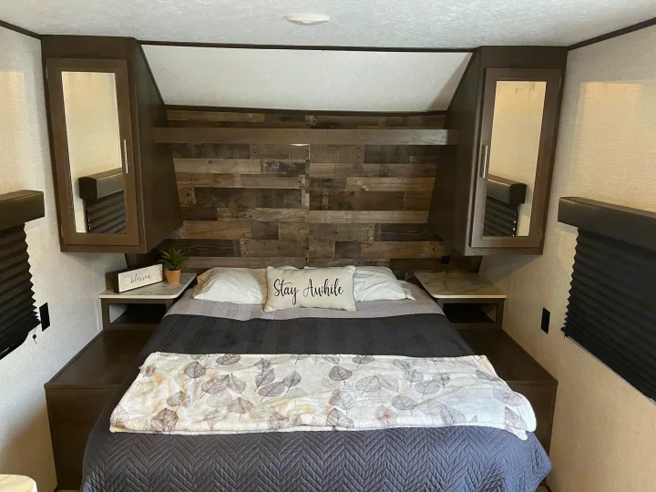 Inside a comfy RV rental bedroom: queen bed, rustic headboard, storage cabinets, and a 'Stay Awhile' pillow.. Dutchmen Coleman Lantern LT 2021