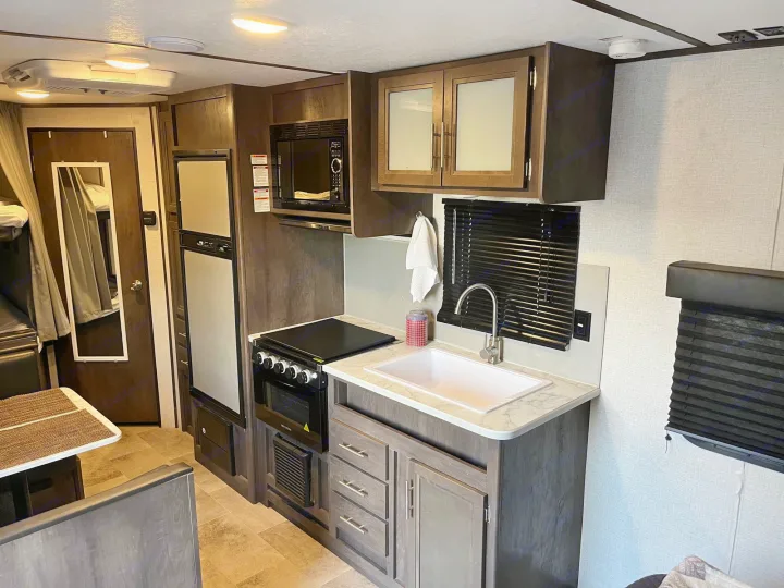 Inside an **rv rental**, showing a compact kitchen area with stove, microwave, sink, and fridge, next to a dining booth.. Dutchmen Coleman Lantern LT 2021