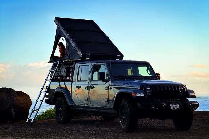 Jeep Gladiator with rooftop tent for an RV rental. A person in the tent enjoys ocean views from this ultimate overlanding setup.. Jeep Gladiator 2022