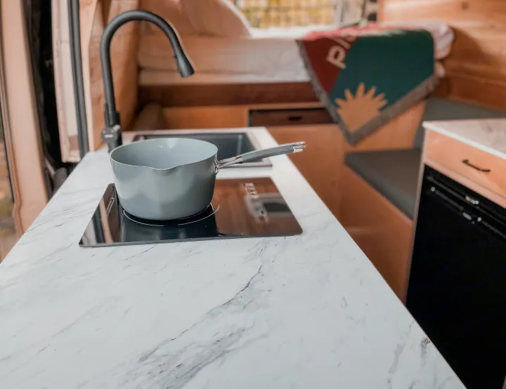 Cozy RV rental interior: a compact kitchen with a saucepan on an induction cooktop, sink, and a comfortable bed in the background.. Ram Promaster 2021
