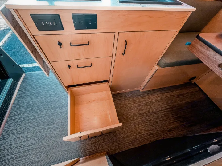 Modern RV rental interior: wooden cabinetry, USB & AC power outlets, and a partially open drawer for storage.. Ram Promaster 2021
