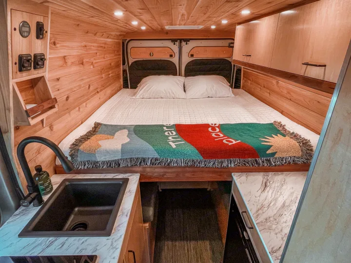 Cozy RV rental interior with a comfortable bed, custom wood paneling, overhead storage, and a functional kitchen sink area.. Ram Promaster 2021