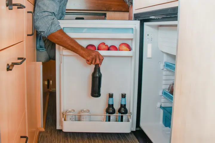 Inside an RV rental, a person takes a bottle from a Dometic fridge, stocked with peaches, bottles, and cans.. Ram Promaster 2021