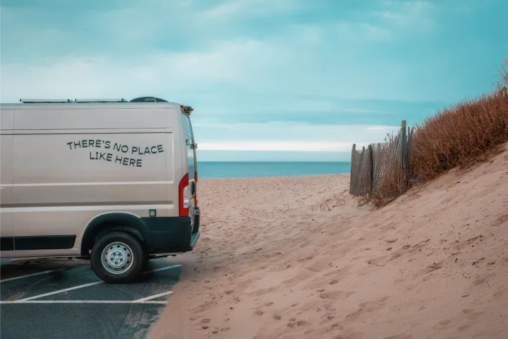 An RV rental van with "THERE'S NO PLACE LIKE HERE" is parked by sandy dunes and a tranquil ocean, embodying a beach getaway.. Ram Promaster 2021
