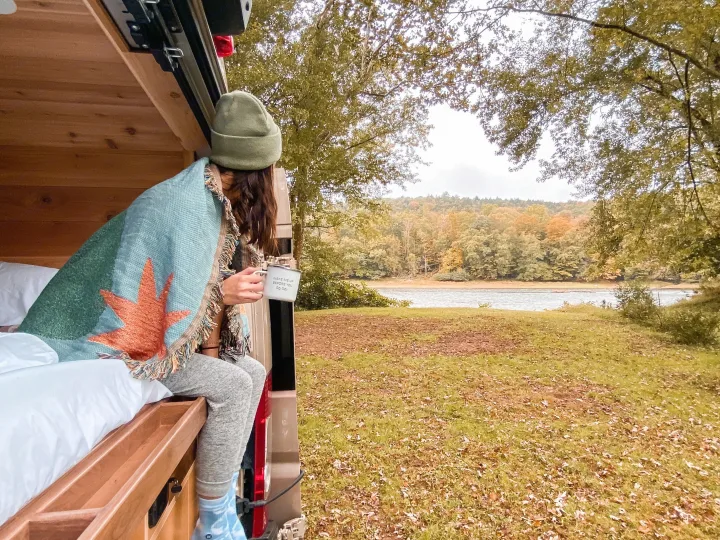 A person wrapped in a blanket sips coffee from an RV rental, enjoying scenic autumn lake views from inside the vehicle.. Ram Promaster 2021