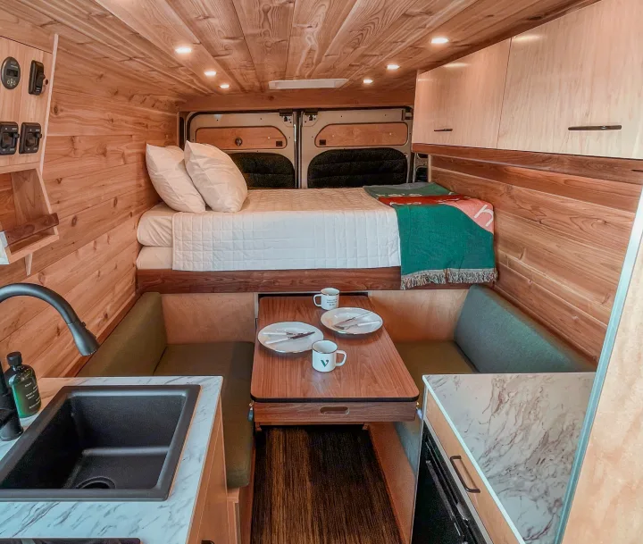 This RV rental's cozy wood interior showcases a raised bed, dining space with benches, and a functional kitchen sink.. Ram Promaster 2021