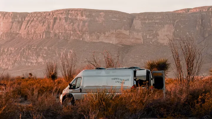 RV rental parked in a sunlit desert setting with towering mountains. The camper van's side reads 'NO PLACE LIKE HERE'.. Ram Promaster 2021