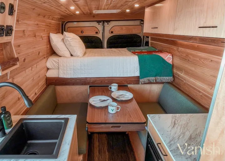 Cozy wood-paneled RV rental interior featuring a comfortable bed, dining table, and compact kitchen. Ideal for road trips.. Ram Promaster 2021