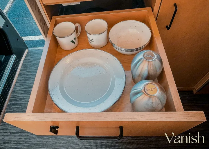 An RV rental kitchen drawer is open, showing organized white enamelware plates, bowls, and mugs, plus two metallic containers.. Ram Promaster 2021