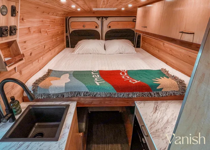 This RV rental interior features a spacious bed, light wood paneling, and a compact kitchen counter with sink.. Ram Promaster 2021