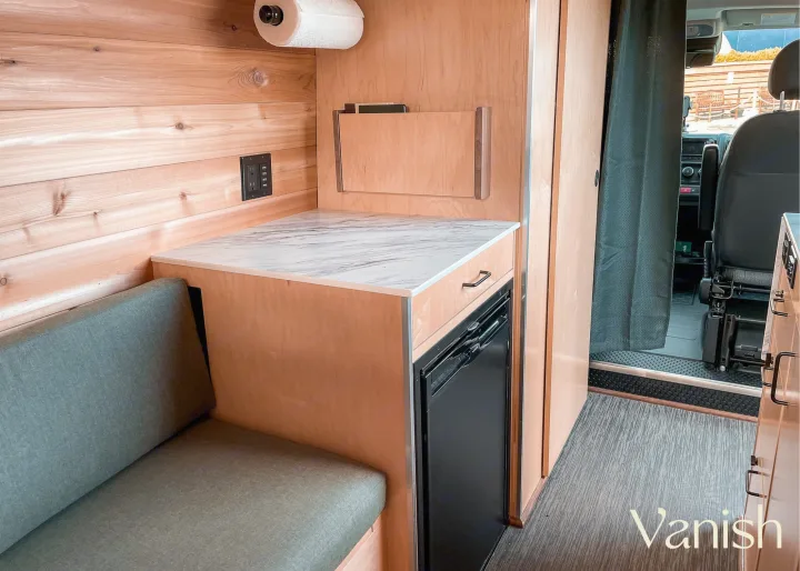 Compact RV rental interior: wood-paneled walls, marble counter, mini-fridge, and comfortable seating.. Ram Promaster 2021