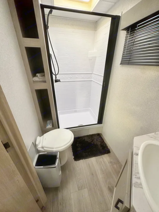 This compact RV rental bathroom features a standing shower with a black frame, toilet, open shelving, and a sink area.. East to West Alta 2022