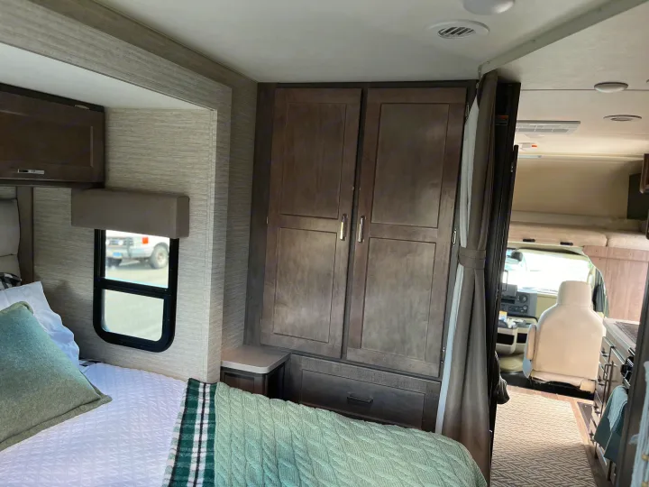 Cozy RV rental bedroom featuring a bed, window, and wood-look wardrobe. View into the front living area.. Winnebago Minnie Winnie 2022