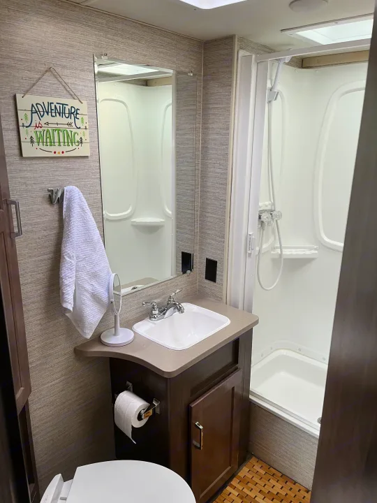 Compact RV rental bathroom featuring a sink, toilet, and shower with a decorative "Adventure is Waiting" sign.. Winnebago Minnie Winnie 2022