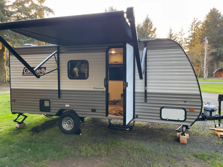 A parked travel trailer RV rental with an extended awning, open door showing the interior, and propane tanks, set for camping.. Coleman 17B 2024