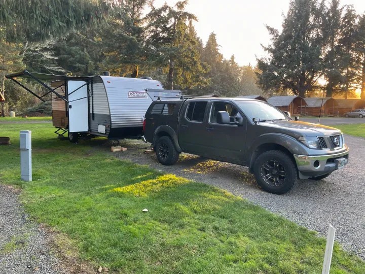 A grey pickup truck tows a Coleman travel trailer, ready for an RV rental adventure in a scenic campground.. Coleman 17B 2024