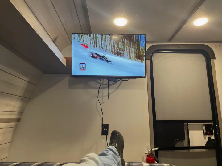Feet up in an RV rental, watching TV showing snowmobiling. Exit door and fire extinguisher visible.. Coleman 17B 2024