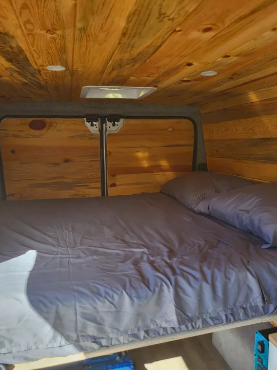 Comfortable RV rental bed with a natural wood interior, featuring bright ceiling lights and storage access.. Dodge Ram Promaster 2021