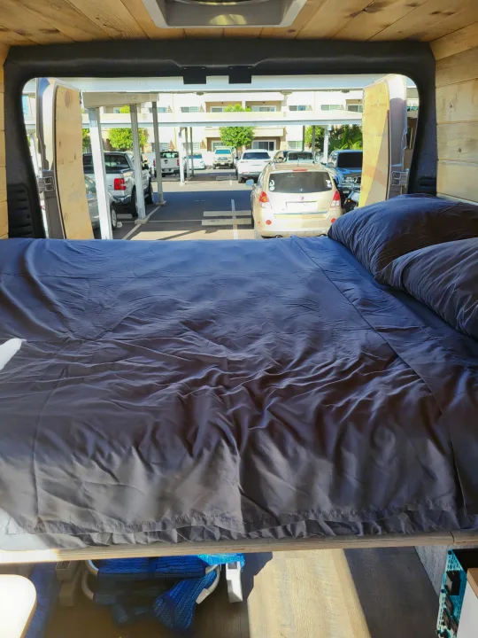 Cozy RV rental interior: a bed with dark bedding, looking out open rear doors to a bright parking lot with cars.. Dodge Ram Promaster 2021