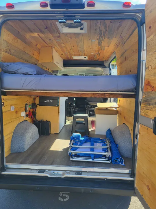 RV rental: Inside a camper van featuring a raised bed, wooden paneling, visible electrical system, and portable seating.. Dodge Ram Promaster 2021