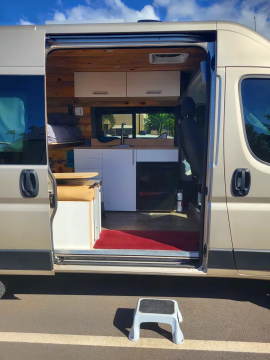 Open side door reveals the well-equipped interior of a tan RV rental van, featuring bed, kitchen, and cabinets.. Dodge Ram Promaster 2021