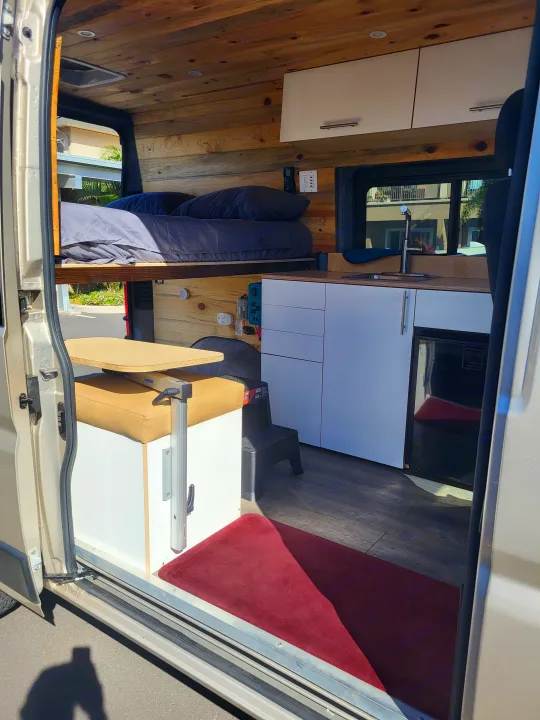 This rv rental shows a custom camper van interior with a raised bed, kitchenette, and versatile table. Ready for your road trip!. Dodge Ram Promaster 2021