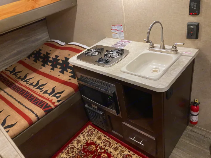 Compact RV rental interior featuring a kitchenette with stove, sink, microwave, and a cozy bunk bed with a patterned blanket.. Forest River Cherokee Wolf Pack Toy Hauler 2017