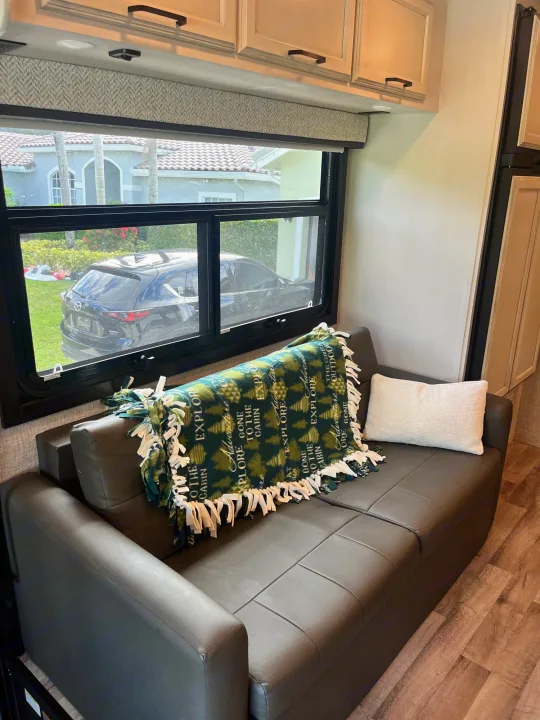 Comfortable RV rental living space with a dark couch, 'Explore' blanket, and window view of an outside house and car.. Entegra Vision 2023