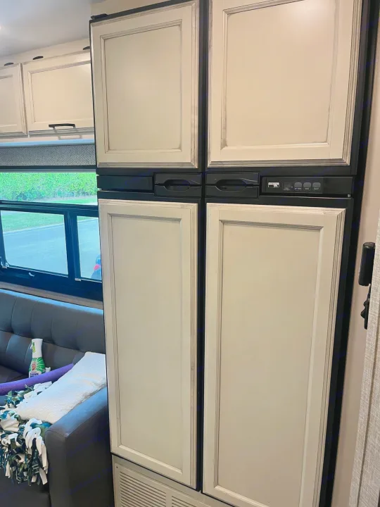 Inside an RV rental: Large paneled refrigerator, upper cabinets, and a living area with a window view.. Entegra Vision 2023