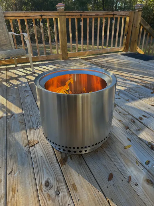 Cozy RV rental deck scene: A stainless steel fire pit with glowing flames on a wooden deck, beside a chair with railing lights.. Grand Design Reflection 2021