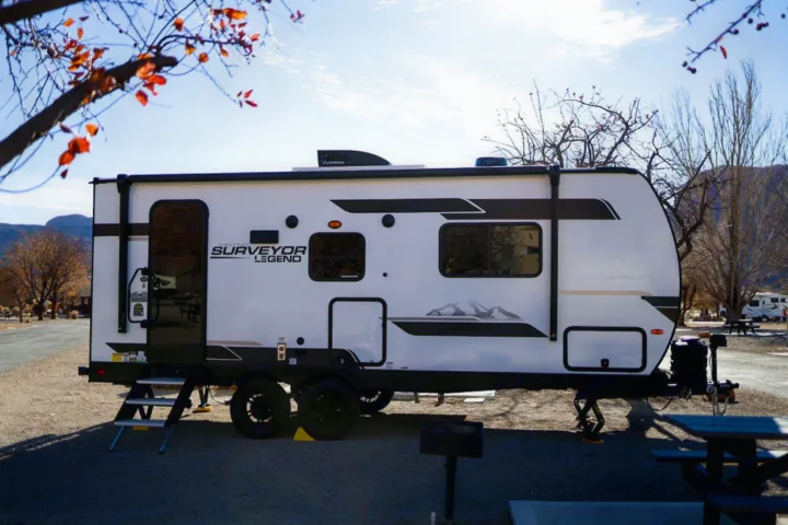A white Surveyor Legend RV rental travel trailer with steps down at a gravel campsite under a blue sky, mountains in back.. Forest River Surveyor Legend 202RBLE 2024