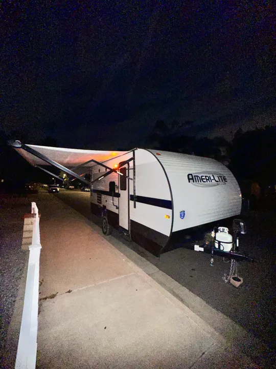 A white Ameri-Lite travel trailer (RV rental) with an extended, lit awning is parked on pavement at night.. Gulf Stream Ameri-Lite Super Lite 2022