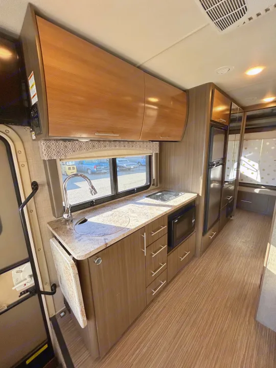 Bright and functional RV rental kitchenette area featuring a sink, cooktop, microwave, refrigerator, and sleek wood cabinets.. Thor Motor Coach Vegas 2018