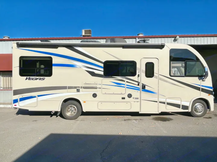 A modern, beige Thor Vegas RV rental with blue and black stripes is parked on asphalt, awning extended.. Thor Motor Coach Vegas 2018