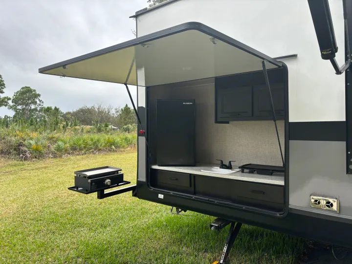 RV rental outdoor kitchen setup with a fridge, sink, and griddle, ready for cooking outdoors in a natural setting.. Jayco Jay Flight 2023